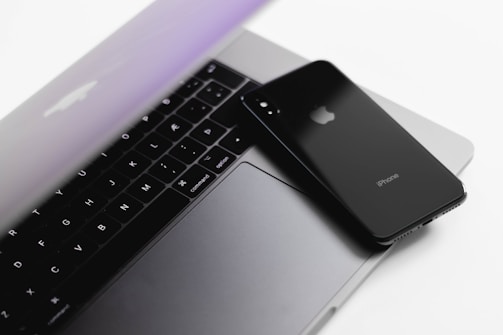A sleek, modern laptop with a partially closed lid is complemented by a black smartphone placed on top of it. The devices sit on a clean, white surface, showcasing minimalistic and contemporary design.