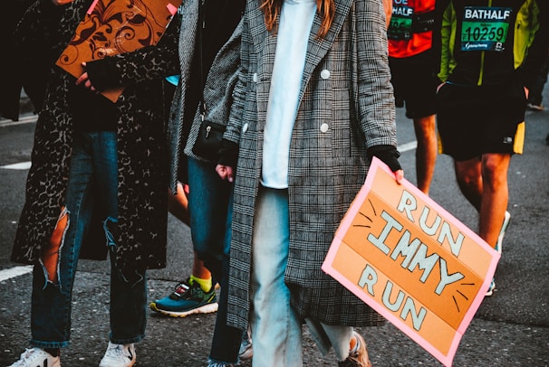 People are walking along a street, one of whom is holding a sign that reads 'Run Immy Run'. The person in the foreground is wearing a long plaid coat and jeans. Other participants in a marathon can be seen in the background, wearing race bibs.