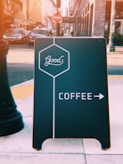 A street view features a black sidewalk sign with the word 'Good' in a hexagon, directing to coffee with an arrow on a busy urban street. In the background, there are parked cars, a 'Do Not Enter' sign, and tall buildings.