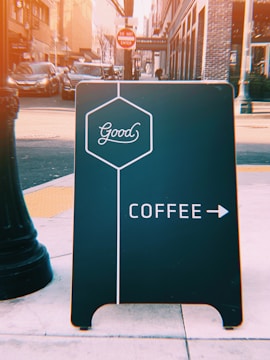 A street view features a black sidewalk sign with the word 'Good' in a hexagon, directing to coffee with an arrow on a busy urban street. In the background, there are parked cars, a 'Do Not Enter' sign, and tall buildings.