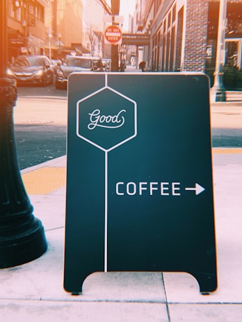 A street view features a black sidewalk sign with the word 'Good' in a hexagon, directing to coffee with an arrow on a busy urban street. In the background, there are parked cars, a 'Do Not Enter' sign, and tall buildings.