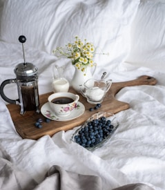 A cozy breakfast scene on a bed with a wooden board hosting a French press coffee maker, a floral teacup filled with coffee, and small pitchers containing cream and sugar. A bowl of fresh blueberries and a small vase of daisies add a natural touch.