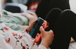 A person is holding a Nintendo Switch console with red and blue Joy-Con controllers. The person is wearing black pants and a white shirt with a floral print. They appear to be sitting or lying down in a relaxed position with another person beside them, partially visible.