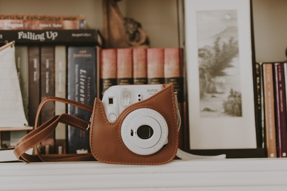 A vintage-style camera is encased in a brown leather case, resting on a white surface. Behind it, books are neatly lined up on a shelf, some visible titles include 'Falling Up' and phrases related to literature. A framed monochrome landscape artwork is seen on the right.