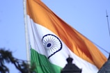 The image depicts the flag of India waving against a clear blue sky. The flag consists of three horizontal stripes of saffron, white, and green, with a blue Ashoka Chakra in the center of the white stripe. Part of the flagpole and a blurred silhouette of an object in the foreground are also visible.