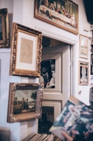 Several framed pictures and posters are arranged on a wall. A classic painting featuring an old scene, likely depicting historical or religious figures, hangs prominently at the top. Below it, numerous frames of varying sizes contain portraits and landscapes. The setting has a vintage and eclectic style, evoking a sense of nostalgia.