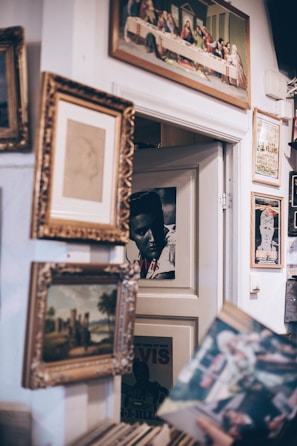 Several framed pictures and posters are arranged on a wall. A classic painting featuring an old scene, likely depicting historical or religious figures, hangs prominently at the top. Below it, numerous frames of varying sizes contain portraits and landscapes. The setting has a vintage and eclectic style, evoking a sense of nostalgia.