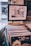 A framed picture hangs on a wall above a collection of vinyl records organized in a container. A sign labeled 'BLUES' is fixed in front of the container, indicating the genre of the records. The framed picture depicts a narrow street or alleyway between buildings.
