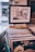 A framed picture hangs on a wall above a collection of vinyl records organized in a container. A sign labeled 'BLUES' is fixed in front of the container, indicating the genre of the records. The framed picture depicts a narrow street or alleyway between buildings.