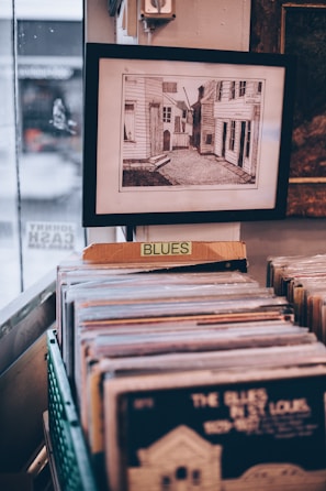 A framed picture hangs on a wall above a collection of vinyl records organized in a container. A sign labeled 'BLUES' is fixed in front of the container, indicating the genre of the records. The framed picture depicts a narrow street or alleyway between buildings.