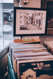 A framed picture hangs on a wall above a collection of vinyl records organized in a container. A sign labeled 'BLUES' is fixed in front of the container, indicating the genre of the records. The framed picture depicts a narrow street or alleyway between buildings.