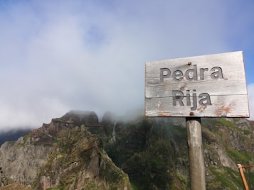 A wooden signpost with the words 'Pedra Rija' stands prominently in the foreground. Behind it, rugged mountains are partially shrouded by mist or clouds, creating a mysterious and serene atmosphere. The mountains are lush with green vegetation, contrasting with the earthy tones of the rocks.