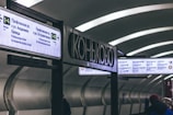 A subway station with signs in both Russian and English showing directions and station information. The station has a modern design with curved ceilings and a dimly lit, gray interior. People are visible walking in the station, wearing winter clothing.