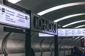 A subway station with signs in both Russian and English showing directions and station information. The station has a modern design with curved ceilings and a dimly lit, gray interior. People are visible walking in the station, wearing winter clothing.