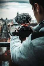 A person wears a grey hoodie while filming or taking photos with a professional camera equipped with a large fluffy microphone. The background is an urban landscape with blurry buildings under a cloudy sky.