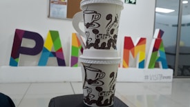 Two stacked disposable coffee cups with brown coffee bean designs are placed in the foreground. In the background, colorful letters spelling 'PANAMA' are visible, with each letter being a different color. The words '#VISITPanama' are written below the letters. The setting appears to be indoors with a tiled floor and a glass window or door on the right.