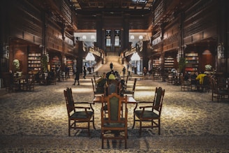An opulent library interior featuring tall, intricately carved wooden bookshelves filled with books. The space is adorned with elegant chandeliers and a grand staircase at its center. Ornate rugs cover the floor, and several people are engaged in reading or conversation at wooden tables throughout the room.