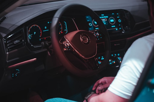 Inside a car, the dashboard is visible with a digital display showing speed and other metrics. The steering wheel is centrally located, with a logo in the middle. The lighting of the dashboard is illuminated in shades of blue and red, creating a modern and slightly futuristic ambiance. A person is seated with their hands resting on their lap, wearing a watch and a light-colored shirt.