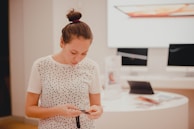 A person with tied back hair is standing in a bright, modern store, focusing on a mobile phone. The individual is wearing a white shirt with black dots. Behind them, there are electronic devices such as tablets and computers on display on a white circular table, along with brochures and other promotional materials.