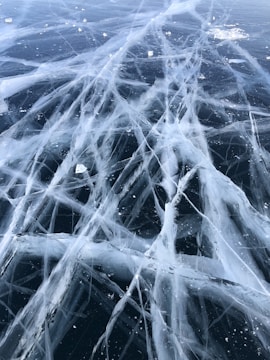 A frozen surface featuring a complex network of white cracks spanning across the ice, creating intricate patterns. Small ice chunks are scattered across the surface.
