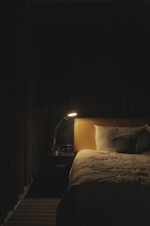 A dimly lit bedroom with a single bed partially illuminated by a small table lamp. The light casts soft shadows, creating a cozy, serene atmosphere. The bed is neatly made, featuring a light-colored duvet and pillows, and a bedside table with a few items placed on it, including the lamp.