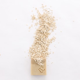 A bar of soap is positioned on a white background with oatmeal flakes scattered over and around it, creating a natural and minimalist setup.