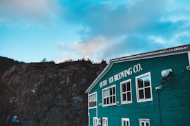 A teal-colored building with the words 'Quidi Vidi Brewing Co.' on its side, located next to a rocky hillside under a partly cloudy sky.