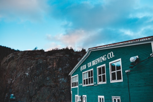 A teal-colored building with the words 'Quidi Vidi Brewing Co.' on its side, located next to a rocky hillside under a partly cloudy sky.