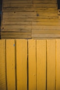 A close-up view of wooden panels painted in a vibrant yellow color. The panels have visible wood grain and knots, emphasizing their natural texture. They are arranged vertically, creating a repeating pattern that fills the frame.