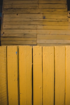 A close-up view of wooden panels painted in a vibrant yellow color. The panels have visible wood grain and knots, emphasizing their natural texture. They are arranged vertically, creating a repeating pattern that fills the frame.