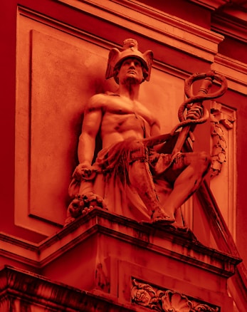 A classical sculpture of a seated male figure, wearing a helmet with wings and holding a caduceus, depicted in a monochromatic red color scheme. The muscular figure is positioned on an ornate pedestal with architectural details in the background.
