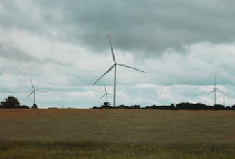 Three wind turbines stand tall in a flat, grassy landscape under a cloudy sky. The blades of the turbines are prominently featured against the backdrop of dark, overcast clouds, suggesting a windy day. The horizon features a mix of grassy fields and distant trees.
