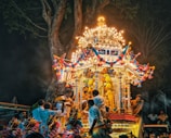 A vibrant and festive temple chariot adorned with bright lights and colorful decorations is surrounded by people in traditional attire participating in a cultural or religious procession under the canopy of a large tree at night.