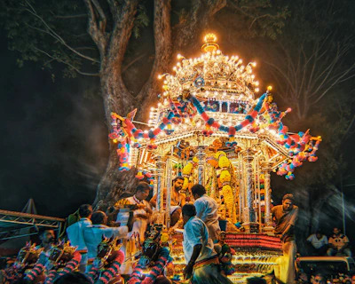 A vibrant and festive temple chariot adorned with bright lights and colorful decorations is surrounded by people in traditional attire participating in a cultural or religious procession under the canopy of a large tree at night.