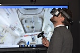 A man wearing a virtual reality headset and holding a controller is engaged in an immersive experience. The background shows a detailed screen of what appears to be the interior of a space station or similar environment, with various equipment and panels visible.