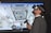 A man wearing a virtual reality headset and holding a controller is engaged in an immersive experience. The background shows a detailed screen of what appears to be the interior of a space station or similar environment, with various equipment and panels visible.