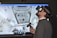 A man wearing a virtual reality headset and holding a controller is engaged in an immersive experience. The background shows a detailed screen of what appears to be the interior of a space station or similar environment, with various equipment and panels visible.