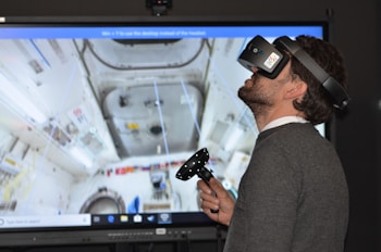 A man wearing a virtual reality headset and holding a controller is engaged in an immersive experience. The background shows a detailed screen of what appears to be the interior of a space station or similar environment, with various equipment and panels visible.
