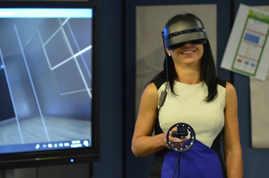 A person is wearing a virtual reality headset and holding a motion controller device, standing in a room with a large screen displaying a virtual environment. The setting suggests a modern, high-tech atmosphere.