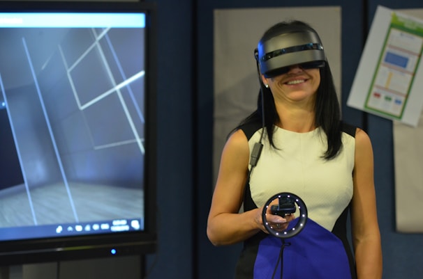 A person is wearing a virtual reality headset and holding a motion controller device, standing in a room with a large screen displaying a virtual environment. The setting suggests a modern, high-tech atmosphere.