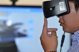 A person is wearing a virtual reality headset, touching the side of the device with their hand as if adjusting it. The background appears blurry, suggesting focus on the individual and the VR device.