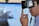 A person is wearing a virtual reality headset, touching the side of the device with their hand as if adjusting it. The background appears blurry, suggesting focus on the individual and the VR device.