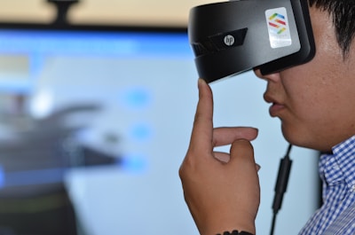 A person is wearing a virtual reality headset, touching the side of the device with their hand as if adjusting it. The background appears blurry, suggesting focus on the individual and the VR device.