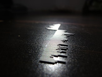 A close-up of a series of small, thin, vertical metal pieces resembling a barcode is depicted on a surface. The lighting creates a sharp shadow, enhancing the contrast between the metallic elements and the dark background.