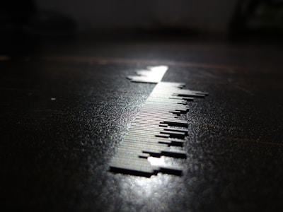 A close-up of a series of small, thin, vertical metal pieces resembling a barcode is depicted on a surface. The lighting creates a sharp shadow, enhancing the contrast between the metallic elements and the dark background.
