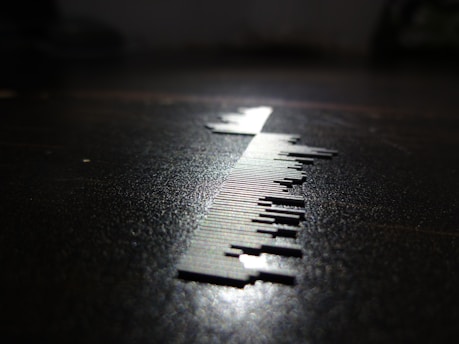 A close-up of a series of small, thin, vertical metal pieces resembling a barcode is depicted on a surface. The lighting creates a sharp shadow, enhancing the contrast between the metallic elements and the dark background.