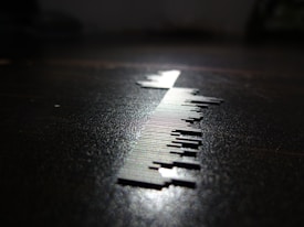 A close-up of a series of small, thin, vertical metal pieces resembling a barcode is depicted on a surface. The lighting creates a sharp shadow, enhancing the contrast between the metallic elements and the dark background.