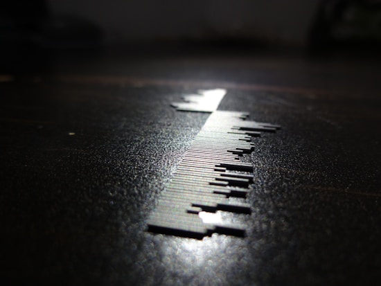 A close-up of a series of small, thin, vertical metal pieces resembling a barcode is depicted on a surface. The lighting creates a sharp shadow, enhancing the contrast between the metallic elements and the dark background.
