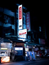A nighttime street view of a building with illuminated signs displaying text in Chinese characters. Several shopfronts with bright lighting are visible, alongside vending machines and street equipment.