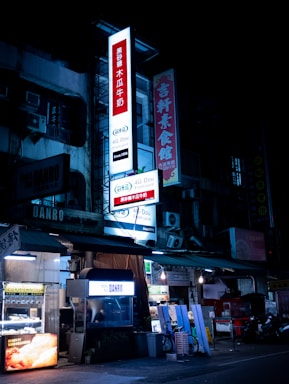 A nighttime street view of a building with illuminated signs displaying text in Chinese characters. Several shopfronts with bright lighting are visible, alongside vending machines and street equipment.